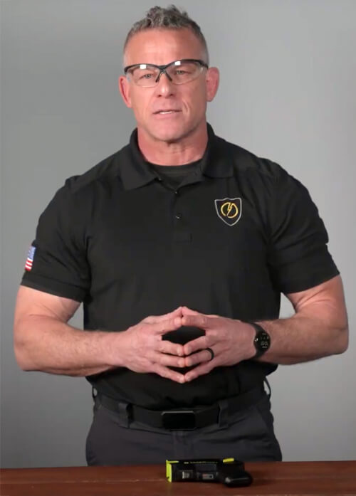 A man standing behind a table wearing safety glasses and has a TASER device on the table 